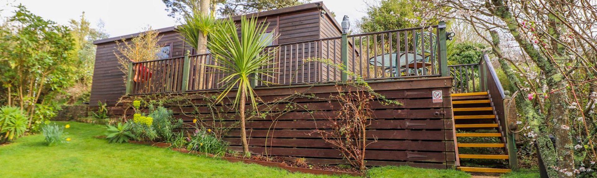 A single-storey timber lodge sits above a raised deck above a large, shrub-lined lawn