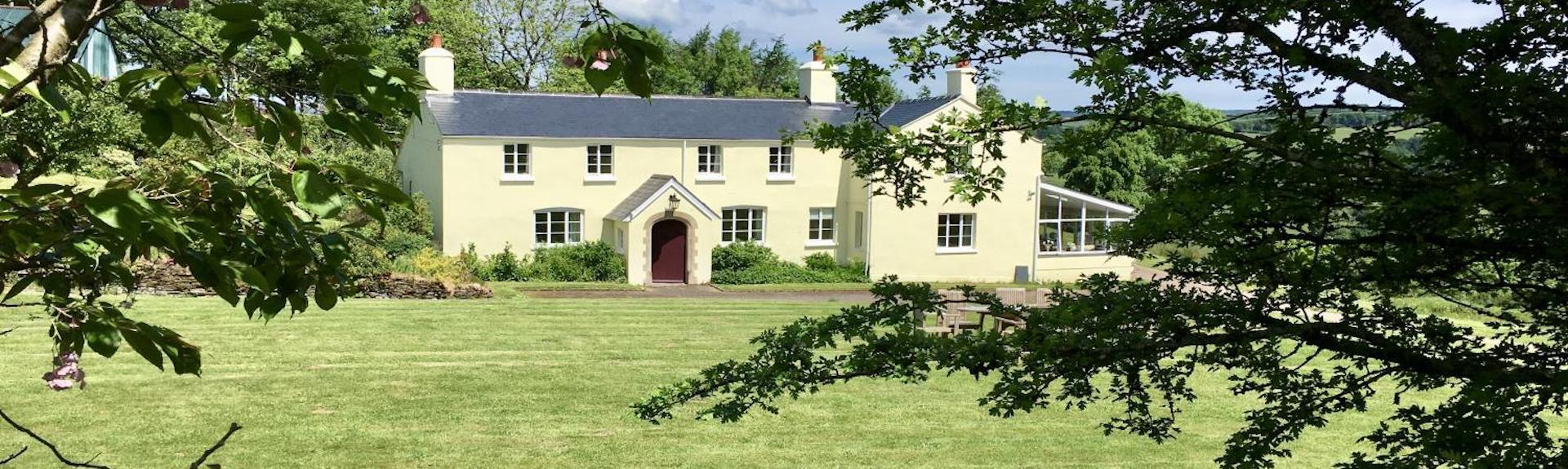 An Exmoor farmhouse overlooks a large lawn surrounded by trees.
