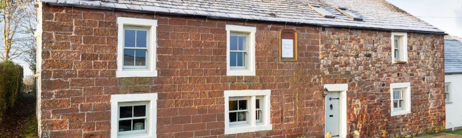 Stone built front exterior of a traditional Cumbrian cottage overlooking a quiet road.