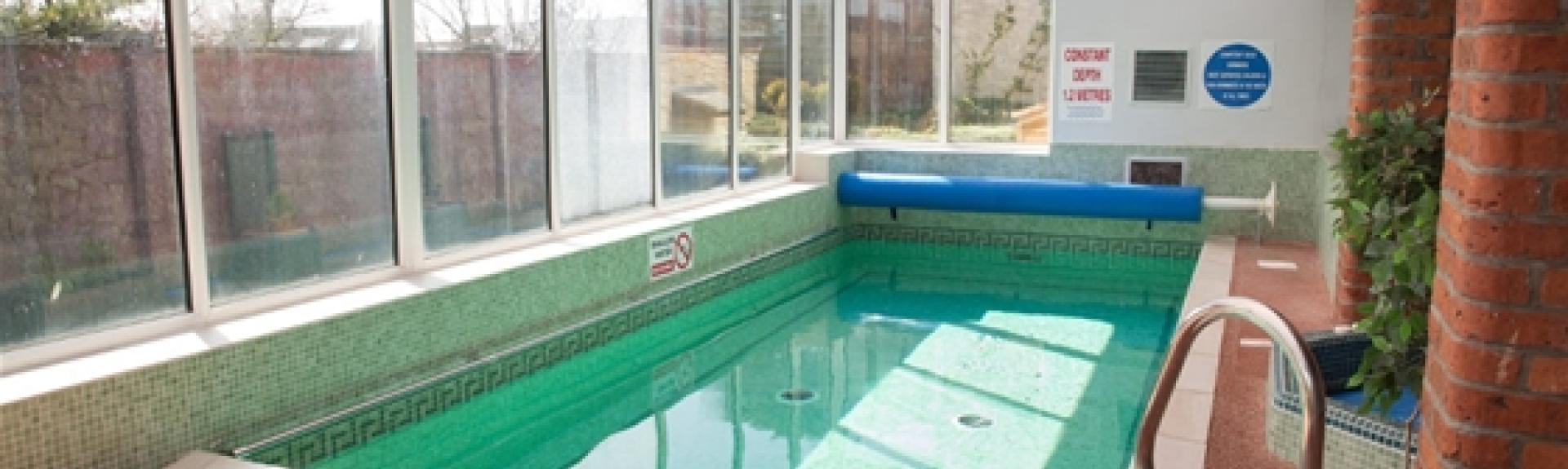 An indoor heated pool with a surrounding terrace. Floor-to-ceiling windows line one side.