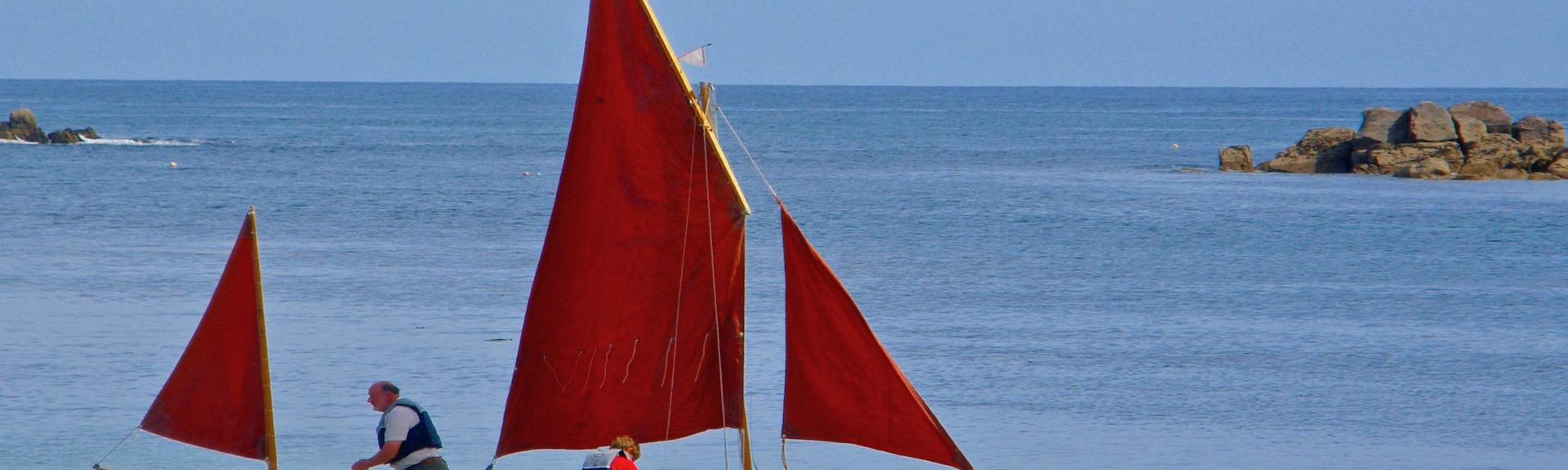 An old-fashioned sailing dinghy sets sail from a sandy beach
