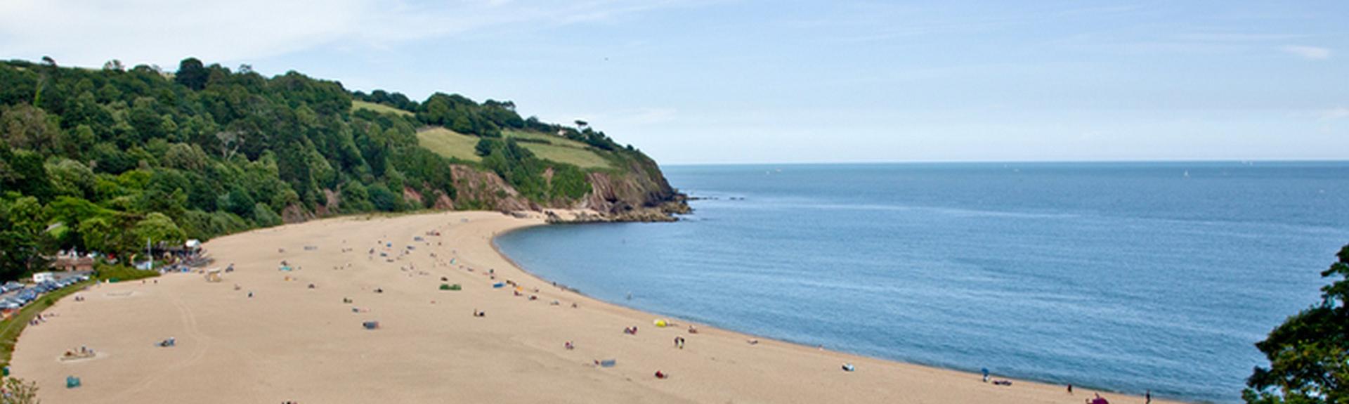 A large sandy cove overlooked by steep-banked wooded hills and cliff tops in South Devon