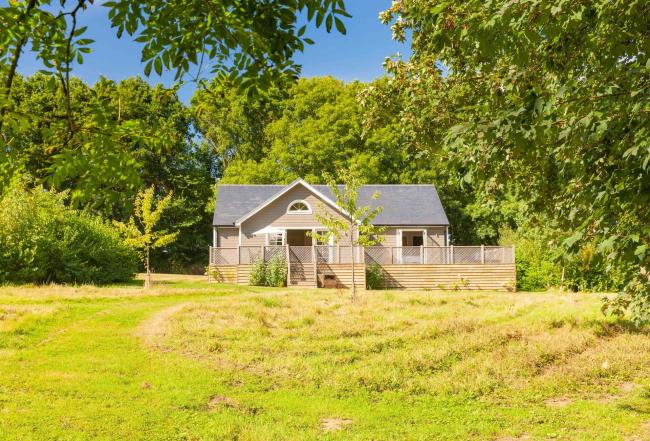 Brook Lodge in the Essex Countryside Outside Colchester. The exerior of a wooden holiday lodge and deck against a sunlit back drop of trees overlooking a field.