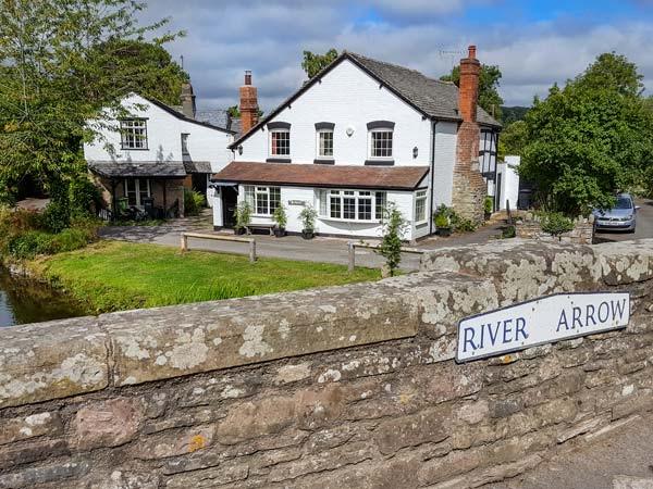 Bridgend Cottage, a Waterside Cottage iin The Banks of The River Arrow in Eardliland. A large 2-storey house with tal chimneys stands behine a lawn which slopes don to a river. In the foregound is a bridge parapit with a sign bearing the name of the river: The River Arrow.