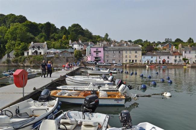 The Quayside at Dittisham on the Dart Estuary A floating pier extends into the River Dart Estuary. Smll boats are moored against the pier. Pastel coloured buildings line the quayside in the background