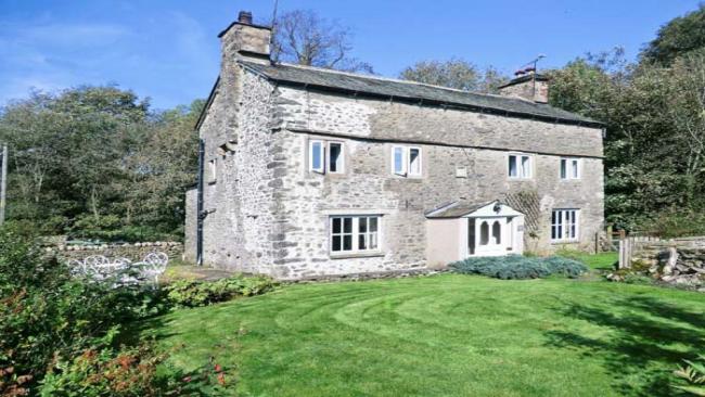 Fleshback Cottage in Kirkby Lonsdale A large double-fronted stone holiday cottage in Kirkby Lonsdale overlooks a newly-mown lawn. Trees and large bushes are to the rear of the house.
