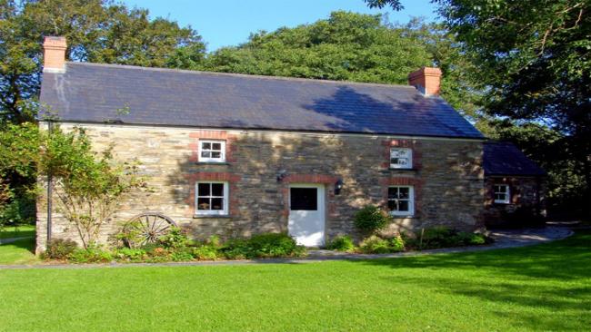 The exterior of  stone-built Welsh farmhouse overlooking  lawn with tall tree to its side and rear. an old cart wheel leans against the wall of the house.