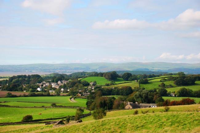 Poto credit: Bridgelands Cottage, Cark-in-Cartmel A Cumbrian landscape view of open countryside in summertime with the houses of the small village of Cark-in-Cartmel half-hidden by trees.