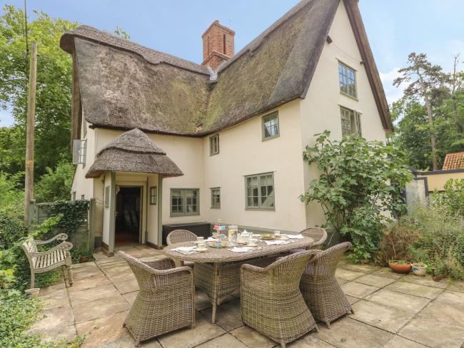 Church Cottage Near Kimbolton in Bedfordshire The extrior of large thatched holiday cottage overlooking a patio garden with whicker picnic table and chairs