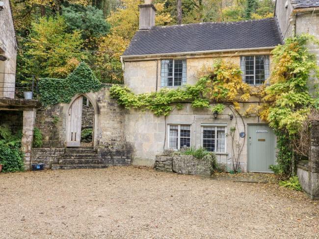 October Cottage in Chalford, Gloucestershire The front of a small Cotswold cottage with quartered windows partially covered by a vine. On one side is an ivy-covered arched entrance to a rear garden.