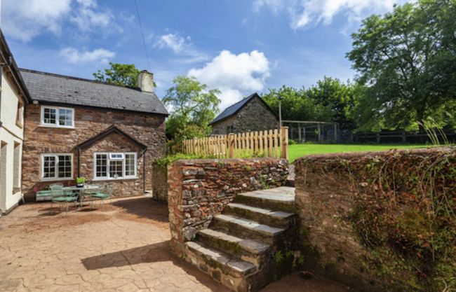 The Cottage at Catford House in Huish Champflower In the background is a 2-storey red-sandstone cottage with outdoor diningtable and chairs on a flagstone patio. I the foreground is a standstone wall with steps leading up to a raised, tree-lined garden