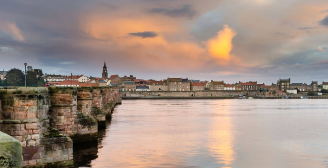 Berwick-upon-Tweed Holiday Cottages A waterside view at sunset in Berwick-upon-Tweed. The stone buttresses of anold bridge lead towards the far bank lined with old 3-storey houses.