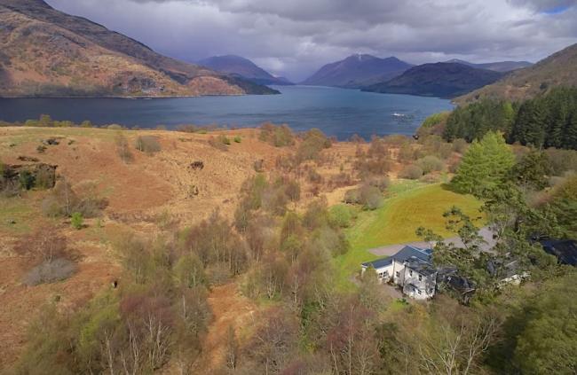 Aerial view of a cottage surrounded by open moorland with a loch in the background.