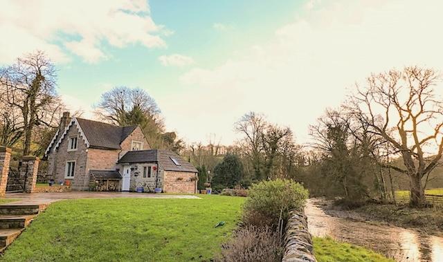 Discover Quality Holiday Cottages To Book in Staffordshire. Photo credit: River Lodge in Leek A river flows past a field and curves round a stone house in the distance.