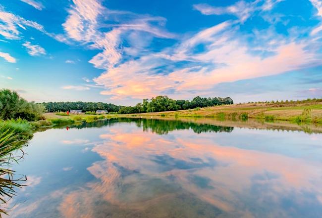 Enjoy Peaceful Rural Retreats in Piddinghoe Two Piddinghoe holiday lodges backed by woods overlook s large lake in which a sunset is reflected.