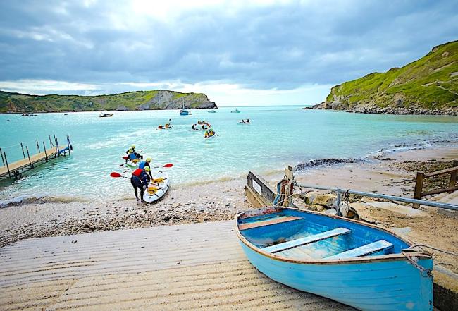 Find Wareham Holiday Cottages Available to Book. Photo credit: Old Nod Cottage Kayaks paddle out to sea from a small Dorset beach on which a small wooden boat is anchored.