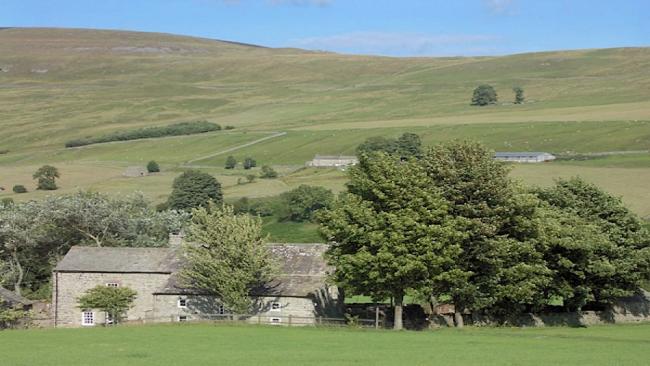 Book a Dales Holiday Home in West Burton A large farmhouse partly surrounded by trees, sits beneath moorland hillside.