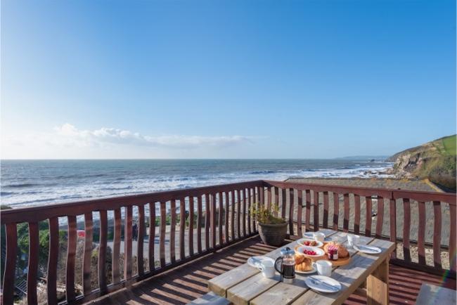 Holiday Cottages in Portwrionkle Beyond a deck with a table laid for a meal, waves crash onto a sandy beach.