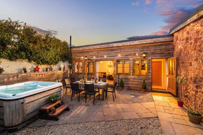 Recharge Your Electric Car When Staying at These Holiday Homes with Their Own Electric Vehicle Chargers A wood-clad single-storey Somerset holiday cottage with a table laid for dinner in a courtyard at dusk.