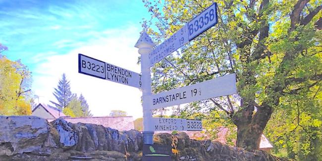 Discover Delightful Exmoor Holiday Cottages in All Directions. An Exmoor signpost against a backdrop of trees with several arms pointing in all directions.
