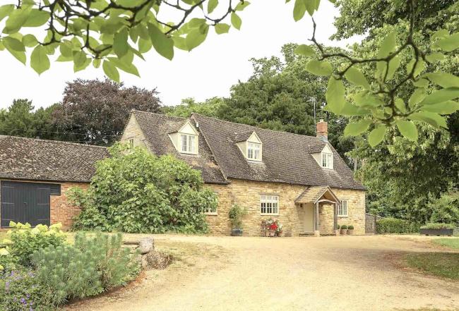 A Cotswold cottage with a backdrop of tall trees sits above a large shingled drive.