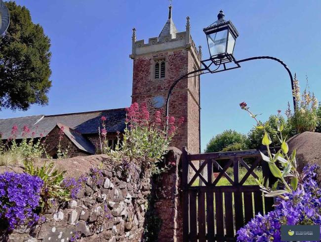 Timberscombe Holiday Cottages lie between Exmoor and The Sea. A church gate with an overhead lantern leads to a red sandstone Somerset church with a tower.