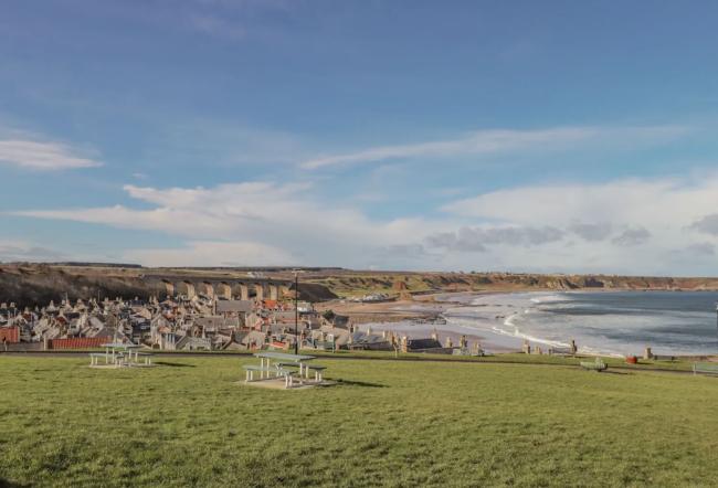 Book 1 Seafield Street in Moray A grass field overlooks a cluster of Moray Firth holiday cottages that face a wave-swept beach curving into the distance.