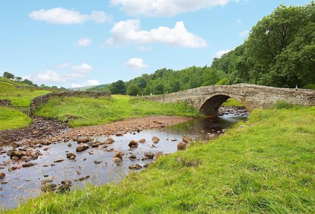 A single arched stone bridge crosses a shallow, rock-filled river in the Yorkshire Dales countryside.