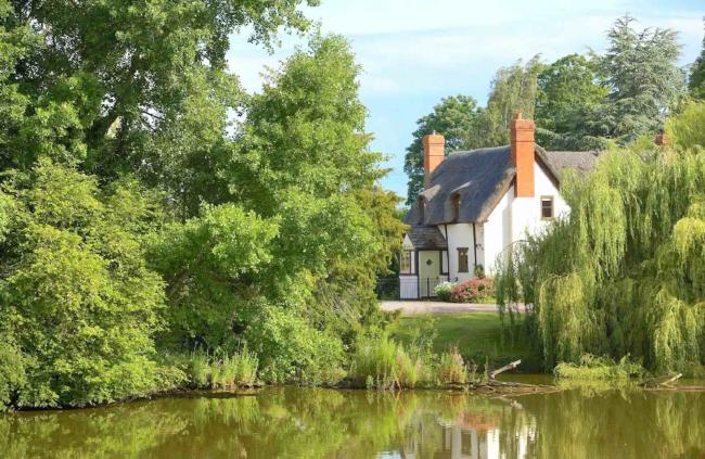A thatched Herefordshire holiday cottage stands between two large willow trees above a lake.