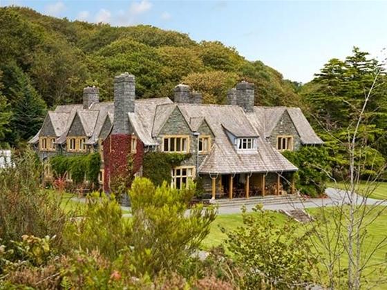 A large slate-roofed house house backed by tall trees overlooks a large lawn.