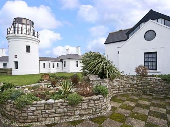 Behind a low stone wall and lawns stands a lighthouse tower and buildings.