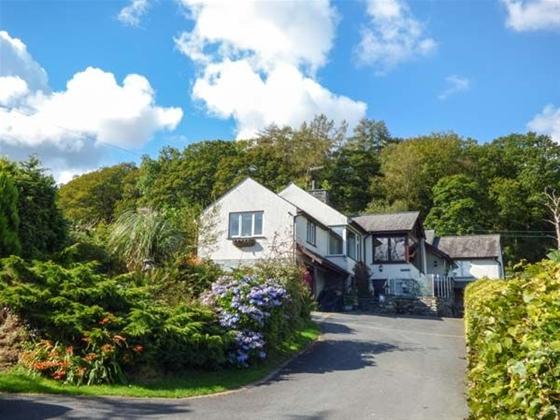 A large Coniston holiday home at the head of a hedge-lined tarmac drive with a mature woodland background.