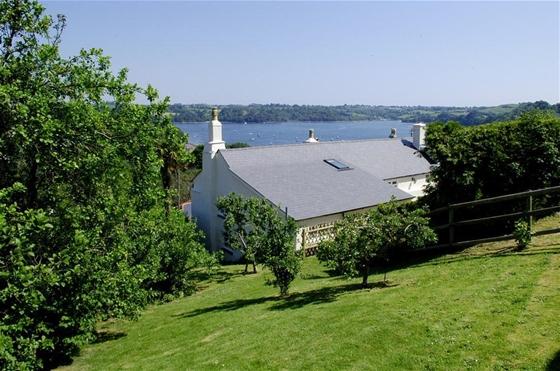 The rooftops of a large holiday cottage surrounded by a sloping lawn and trees with a river estuary in the distance.