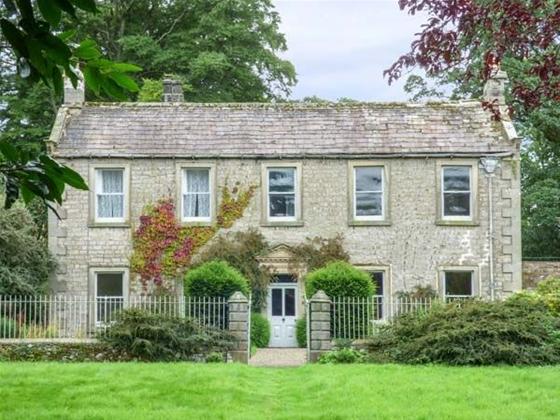A classic country house with a row of five windows overlooks a shrub0filled garden lined by wrought iron railings. 
