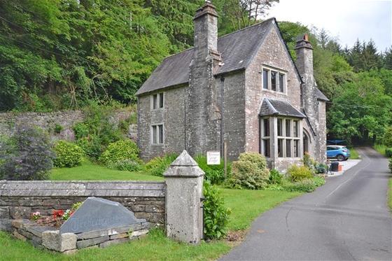 A drive leds to a tall stone house with a mullioned bay window. A wood grows behind the house.