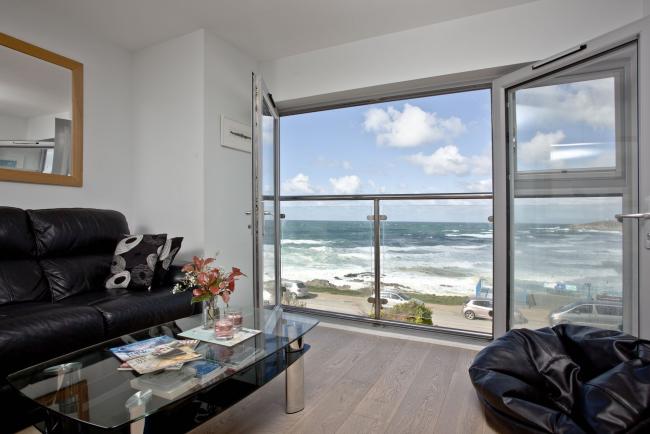 A holiday apartment lounge with floor to ceiling windows beyond which is a view of rhe surf on Ffistral Beach