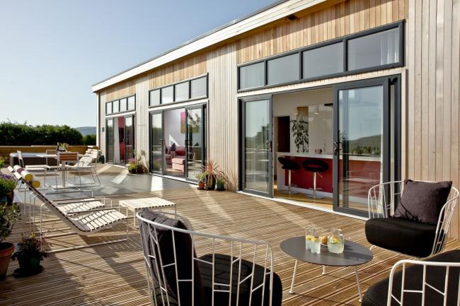 Exterior of a contemporary eco-lodge with large floor-to-ceiling plate glass windows and doors opening onto a wooden deck with a hot tub.