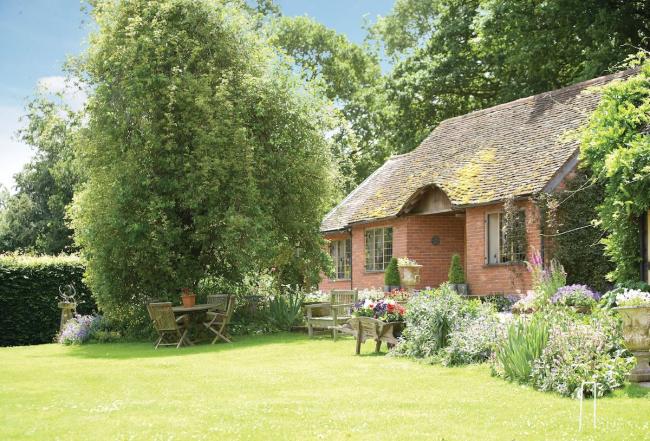 Exterior of s brick-built period garden cottage backed by mature woodland and in front of a lawn with flower-filled borders.
