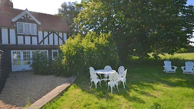 Outdoor dining chairs cluster around a table on a tree-lined lawn on Osea Island. Behind them is a cross-timbered holiday cottage on Osea Island.