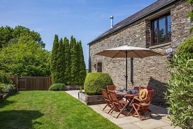 A table  with parasol and chairs tands on a sunny patio behind a stone built holiday cottage in Devon.