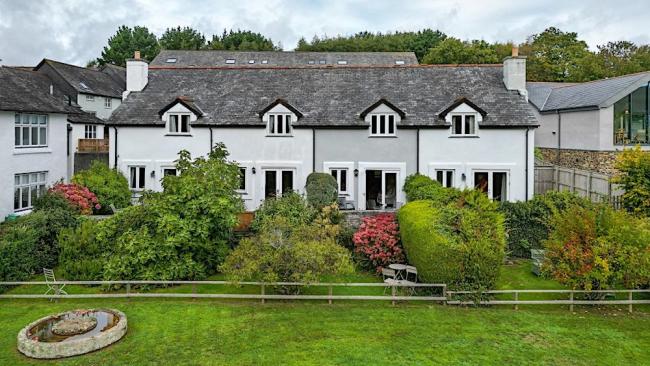 A terrace of 4 slate-roofed cottages over looking a lawn with tall shrubs and an open field in Dittisham. 