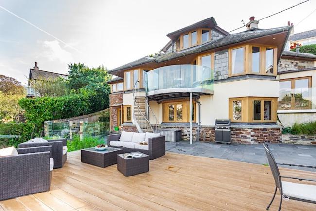 A wooden deck with lounge furniture sits in front of a contemporary 3-storey waterside holiday home.
