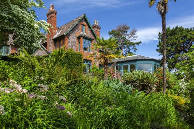 A House with large conservatory is surrounded by a garden with lawns and mature shrubs.