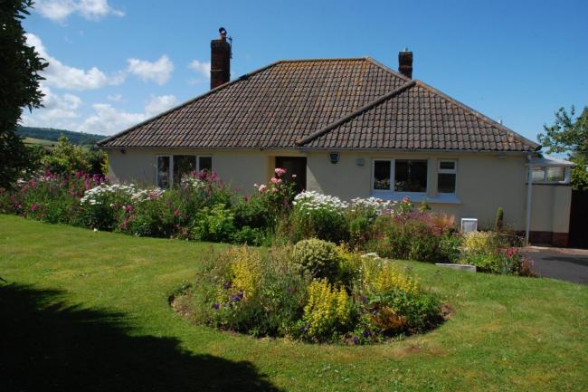 A bungalow sits behind a large lawn with flower-filled borders.
