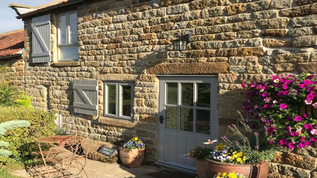 Exterior of a sandstone cottage with a courtyard lined by flowering shrubs.
