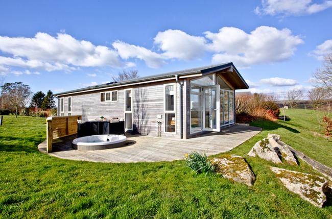 A wooden eco-lodge with a hot tub stands in an open field.