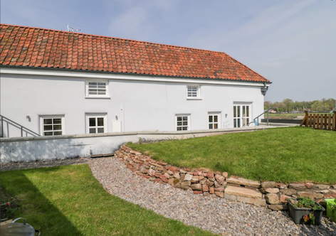 A rural holiday cottage with a terracotta roof sits behind a lawn with a shingled footpath.