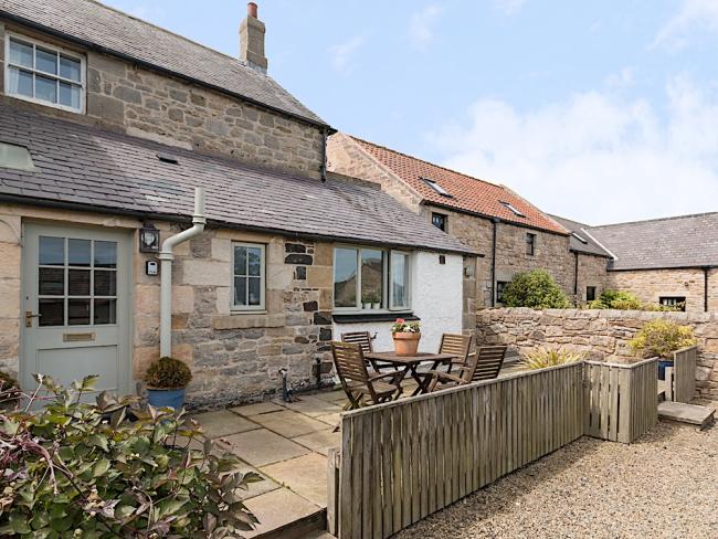 Exterior of a stone-built holiday cottage with ground fllor extension behind a paveed  garden with dining table and chairs.