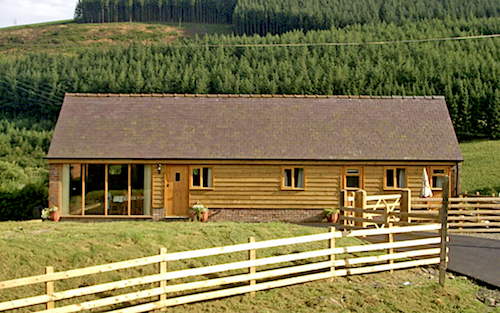Exterior of a wooden eco-lodge with woods to the rear and open fields in the foreground.