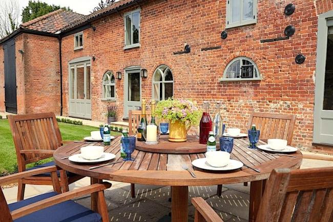 A wood dining table set for breakfast stands on a courtyard in front of a brick-built Norfolk cottage with arched windows.